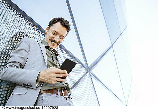 Smiling businessman using smart phone in front of modern glass building