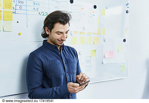 Smiling businessman using smart phone in front of whiteboard at office