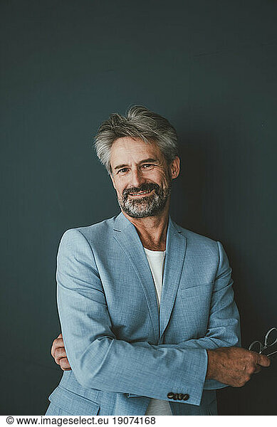 Smiling businessman with arms crossed leaning on teal background