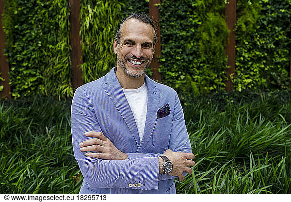 Smiling businessman with arms crossed standing in front of hedge