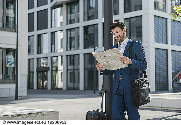 Smiling businessman with baggage reading newspaper in the city
