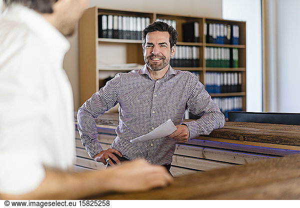 Smiling businessman with document looking at colleague in wooden open-plan office