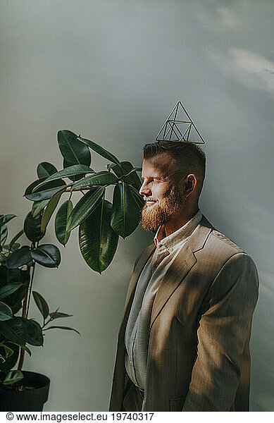 Smiling businessman with eyes closed balancing metal pyramid on head in front of wall