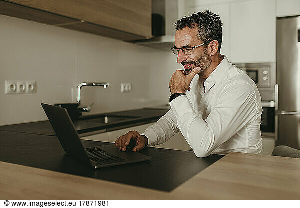 Smiling businessman with hand on chin using laptop at home