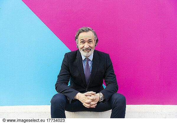 Smiling businessman with hands clasped sitting in front of wall