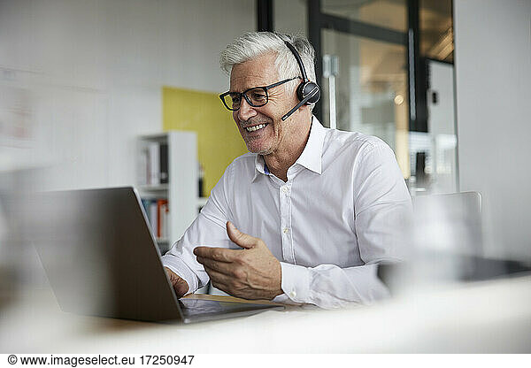 Smiling businessman with headset gesturing while talking to video call on laptop in office