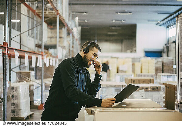 Smiling businessman with headset holding document at distribution warehouse
