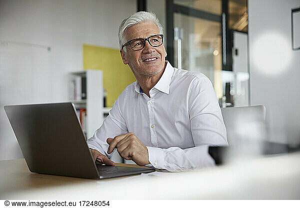 Smiling businessman with laptop contemplating in office