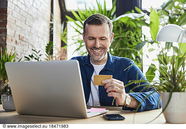 Smiling businessman with laptop holding credit card at loft office