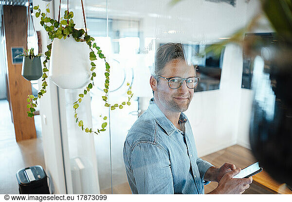 Smiling businessman with smart phone seen through glass wall in office