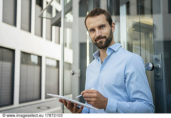Smiling businessman with tablet PC standing in front of glass