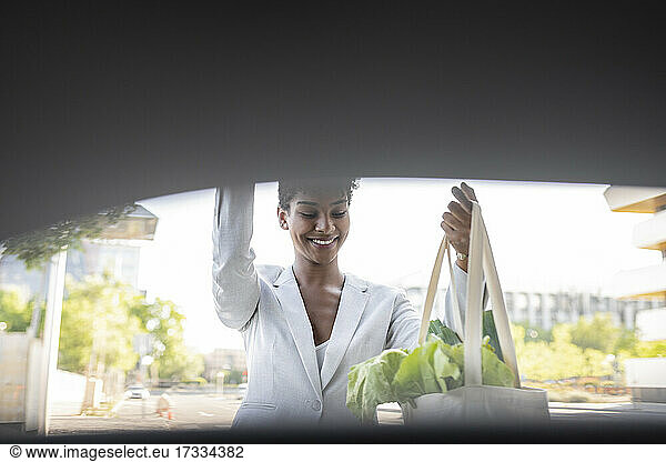 Smiling businesswoman closing car trunk while holding bag of vegetables