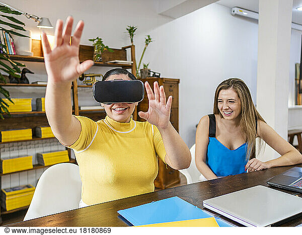 Smiling businesswoman gesturing with VR goggles sitting by friend at table