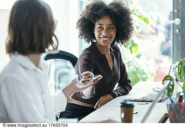 Smiling businesswoman giving mobile phone to colleague at desk in office