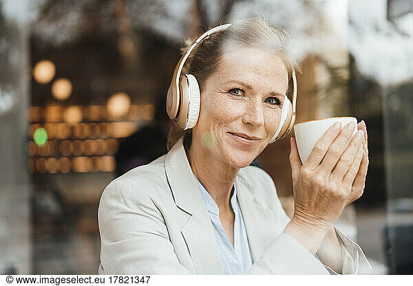 Smiling businesswoman holding coffee cup enjoying music listening through wireless headphones in cafe