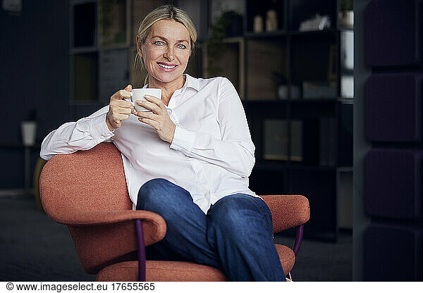 Smiling businesswoman holding coffee cup sitting on chair in office