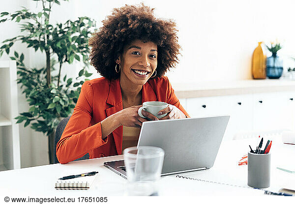 Smiling businesswoman holding coffee cup sitting with laptop at desk