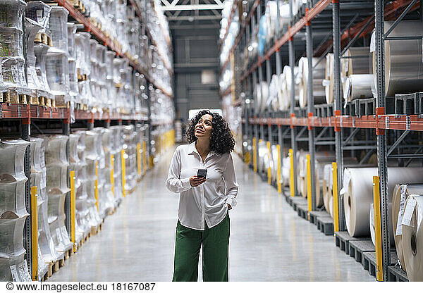 Smiling businesswoman holding smart phone standing with hand in pocket at factory