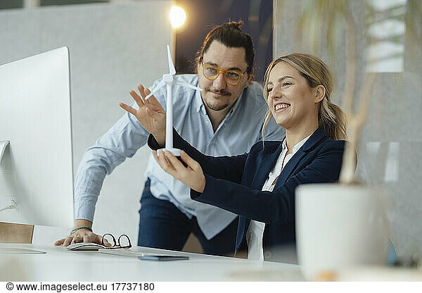 Smiling businesswoman holding wind turbine model discussing with colleague in office