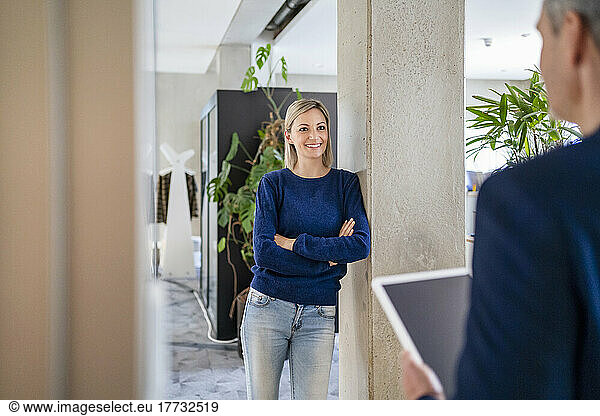Smiling businesswoman leaning against column in office looking at colleague