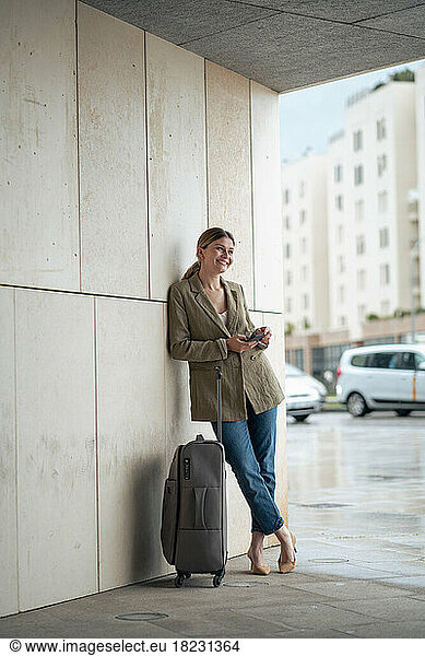 Smiling businesswoman leaning by luggage on wall