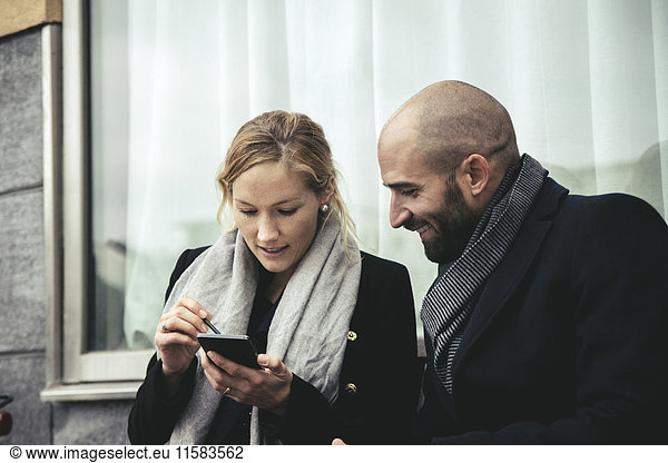 Smiling businesswoman looking at businesswoman using smart phone against window