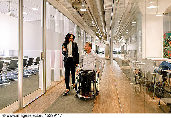 Smiling businesswoman looking at disabled colleague sitting on wheelchair in corridor at work place