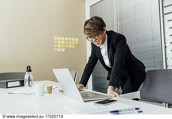 Smiling businesswoman looking at laptop while leaning at desk in office