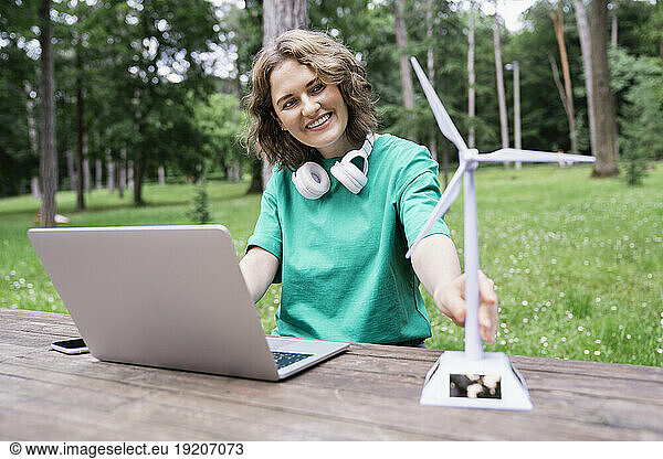 Smiling businesswoman looking at wind turbine model with laptop on table in forest