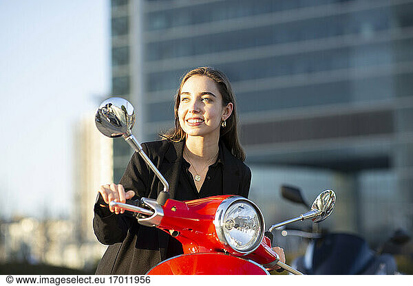 Smiling businesswoman looking away while sitting on motor scooter during sunset