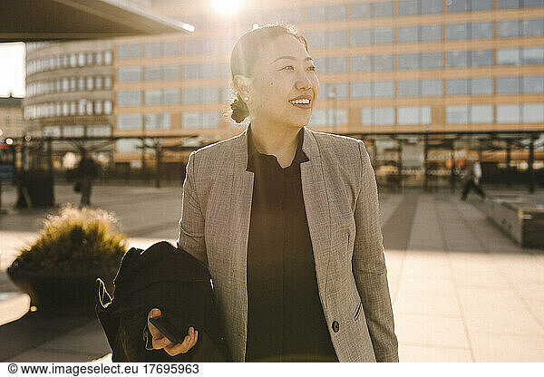 Smiling businesswoman looking away while standing with overcoat on sunny day