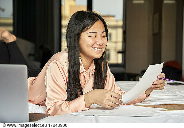 Smiling businesswoman reading document while lying on bed at hotel