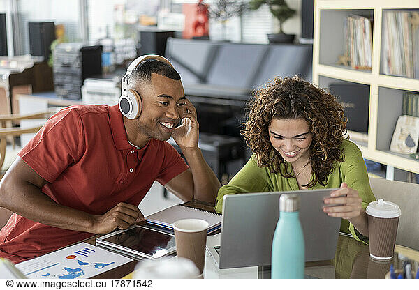 Smiling businesswoman sharing laptop with colleague wearing wireless headphones in office
