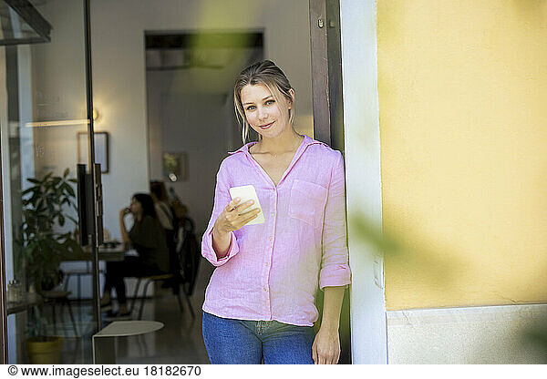 Smiling businesswoman standing by wall with smart phone at cafe