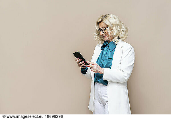 Smiling businesswoman surfing net through smart phone against beige background
