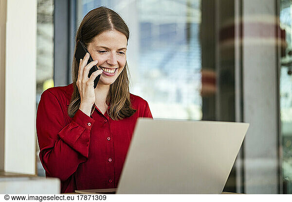Smiling businesswoman talking on smart phone and using laptop in office