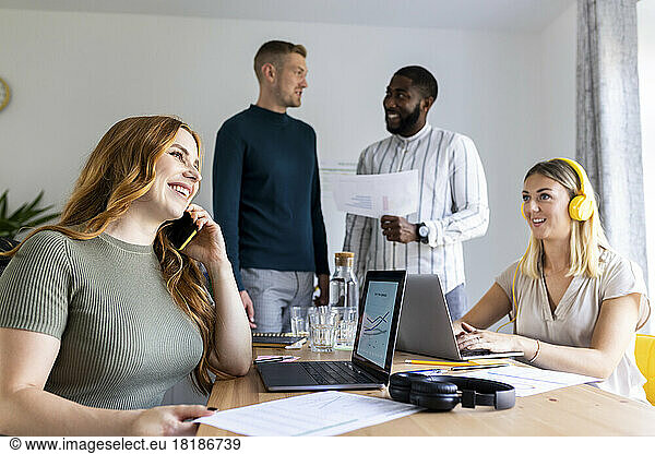 Smiling businesswoman talking on smart phone at desk by colleagues in office