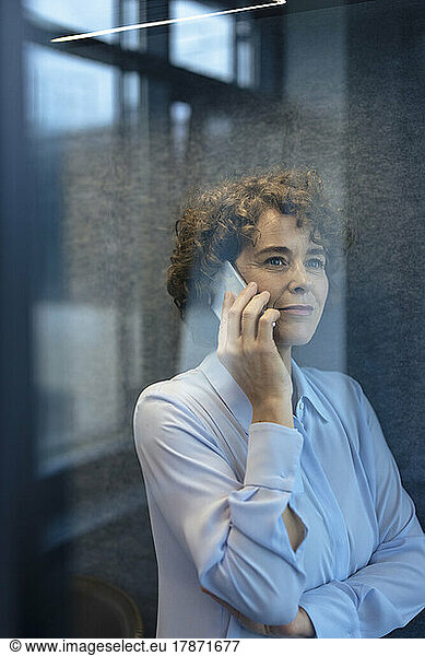 Smiling businesswoman talking on smart phone seen through glass
