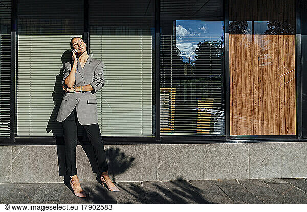 Smiling businesswoman talking through smart phone in front of glass wall on sunny day