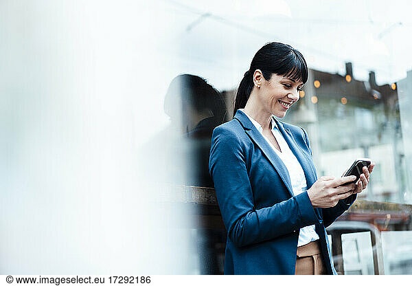 Smiling businesswoman using mobile phone while leaning on cafe window