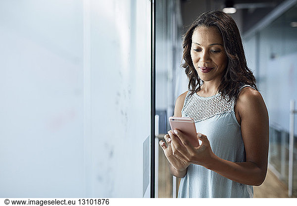 Smiling businesswoman using mobile phone while standing by glass wall at creative office