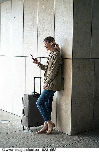 Smiling businesswoman using smart phone by luggage leaning on wall in tunnel