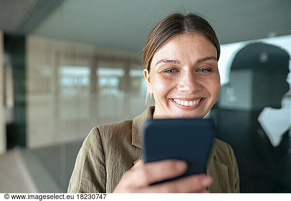 Smiling businesswoman using smart phone leaning on glass wall