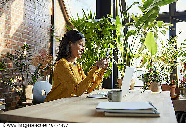 Smiling businesswoman using smart phone sitting at desk in loft office