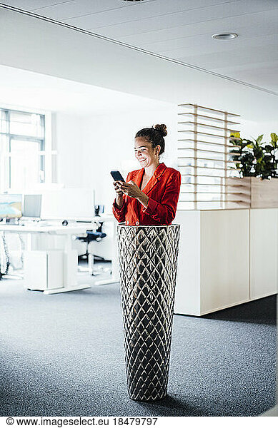 Smiling businesswoman using smart phone standing inside of planter at office