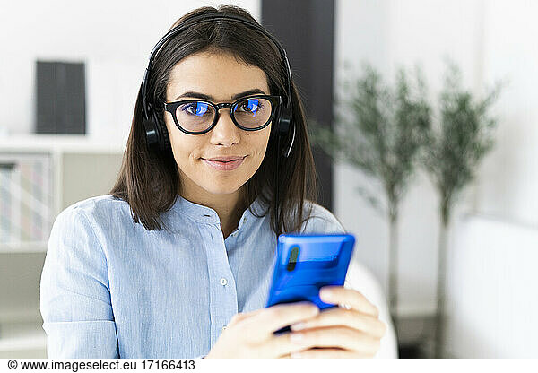Smiling businesswoman wearing eyeglasses and microphone headset using mobile phone while sitting at office