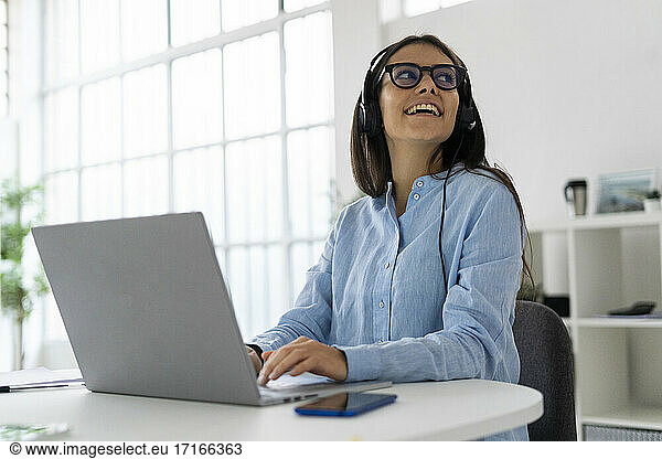Smiling businesswoman wearing microphone headset looking away while sitting with laptop in office