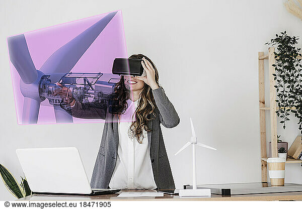 Smiling businesswoman wearing VR goggles and touching wind turbine hologram
