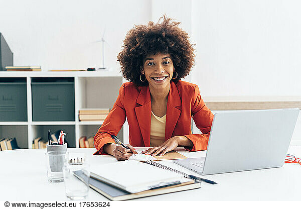 Smiling businesswoman with Afro hairstyle sitting with laptop at desk in office