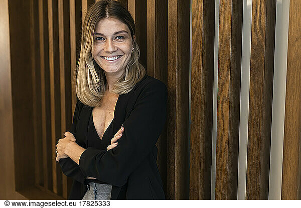 Smiling businesswoman with arms crossed in front of wooden wall at hotel